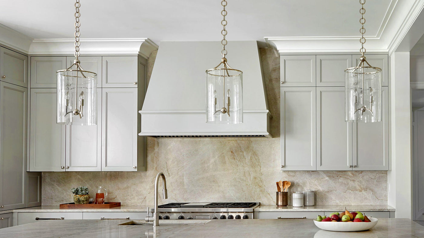 A trio of Minerva Lanterns, featuring discrete polised nickel fittings and blown glass cylinders, designed by Robert A.M. Stern Archtiects for Remains Lighting Co., hanging in a kitchen with muted gray cabinets and marble counter and walls.