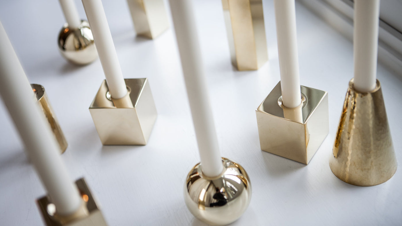 An array of Remains Lighting Co. candlesticks, in round, square and triangular shapes and finishes ranging from polished to peened brass, sit on a white table in the natural light, viewed from above.