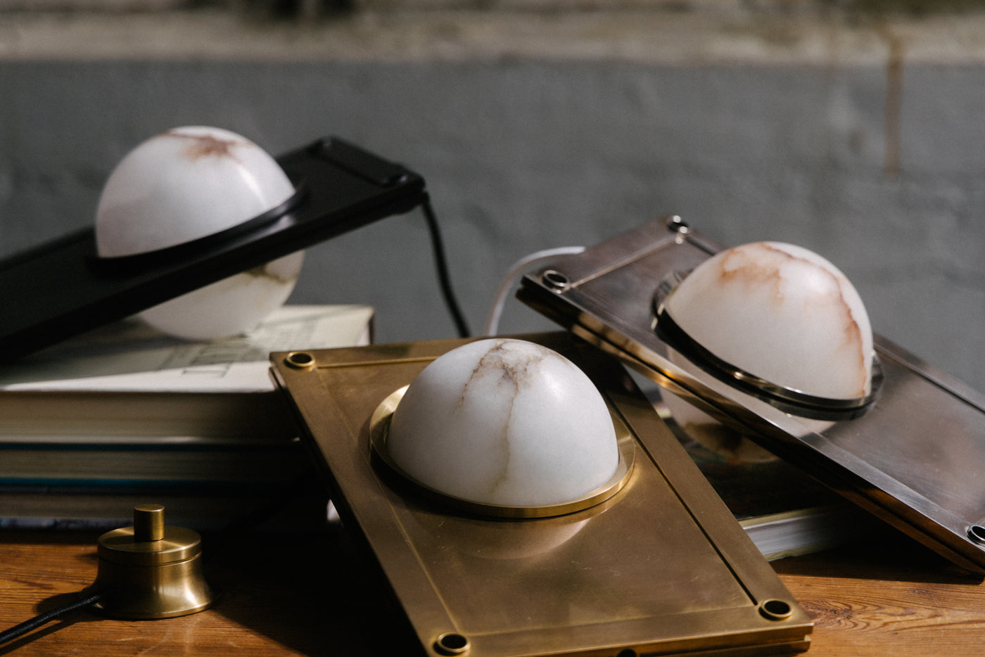 Three Nima Table Lights - consisting of a white alabaster globe set, porthole-style, within a refined slab of bronzed, antiqued, or nickeled brass - are arrayed at various angles on a wooden table with books.