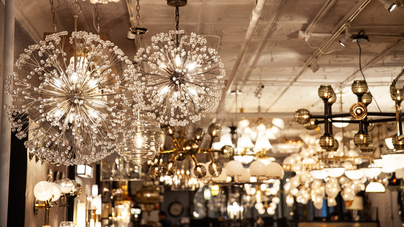 A view of the Remains Showroom ceiling filled with warmly illuminated lights fixtures. Two Dandelion Chandeliers on the left and a Haussmann Hex Chandelier with Beehive Glass.