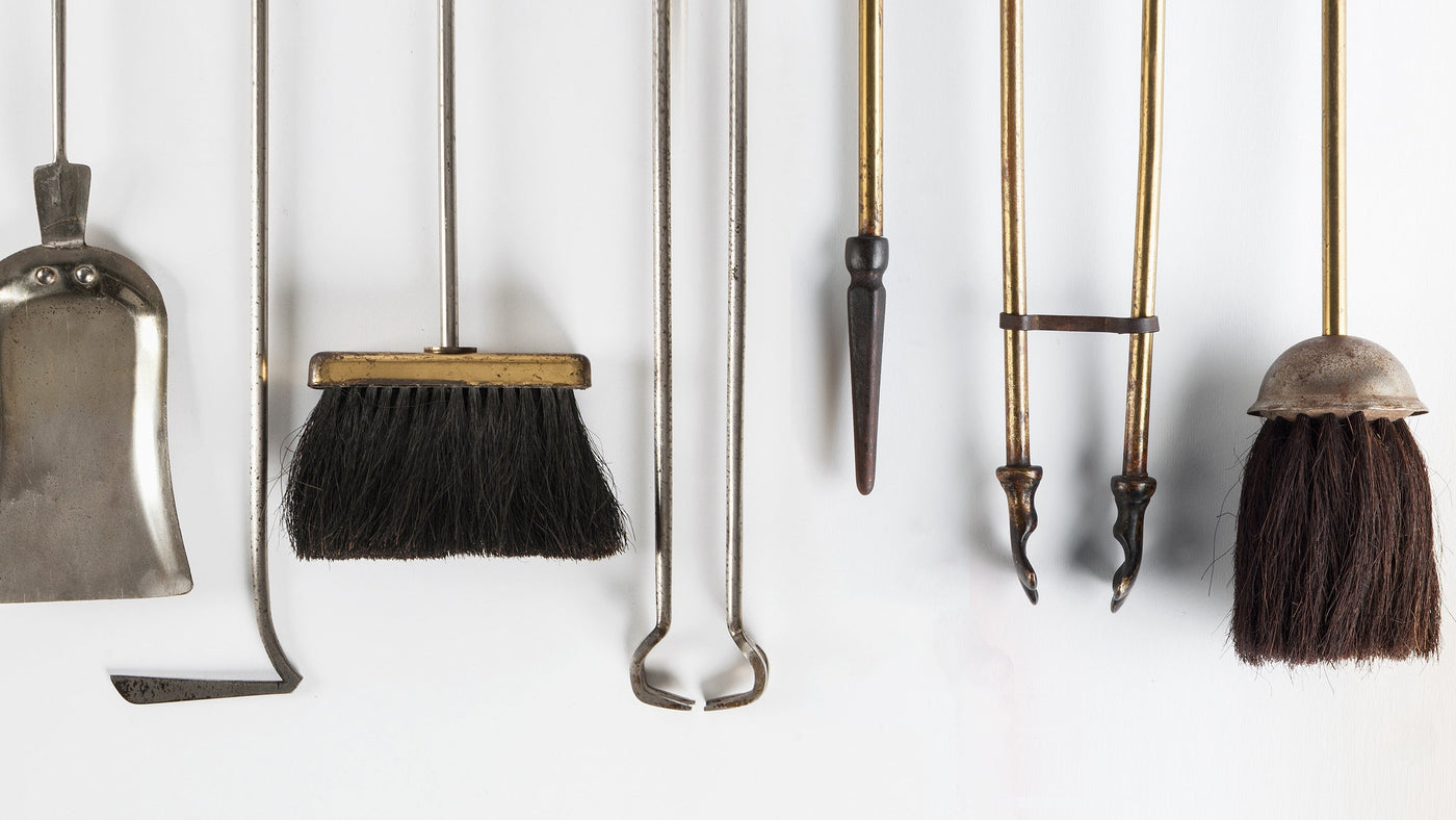 An assortment of vintage and antique fireplace tools from the Remains Lighting Co. Fireplace Collection, including aged brass tongs, cast iron pokers, a nickel shovel, and brushes with brass handles.