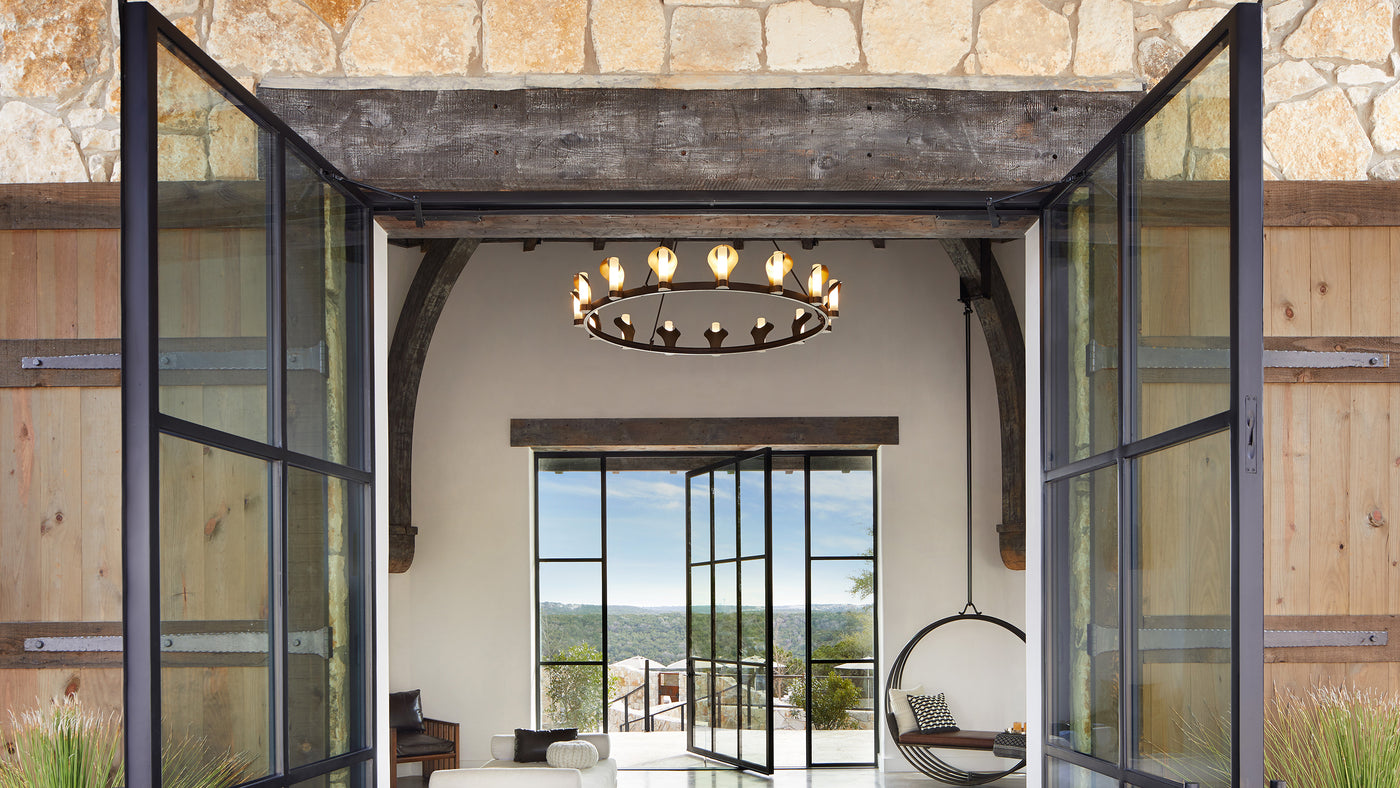 A large scale dark bronze ring chandelier lit by 15 evenly spaced brass lanterns with glass cylinder shades hangs in a white vaulted space with floor to ceiling windowed doors leading out to a patio and view of the hilly wooded landscape in the distance.
