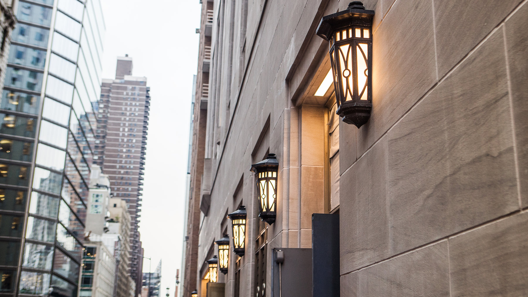 A group of traditional bronze and opal glass exterior wall sconces shown lit along the limestone wall of a New York City residential building on Central Park South