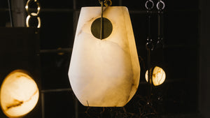 A white basket-shaped alabaster lantern with brass accents, suspended by leather-wrapped hooks, hangs in a darkened room.