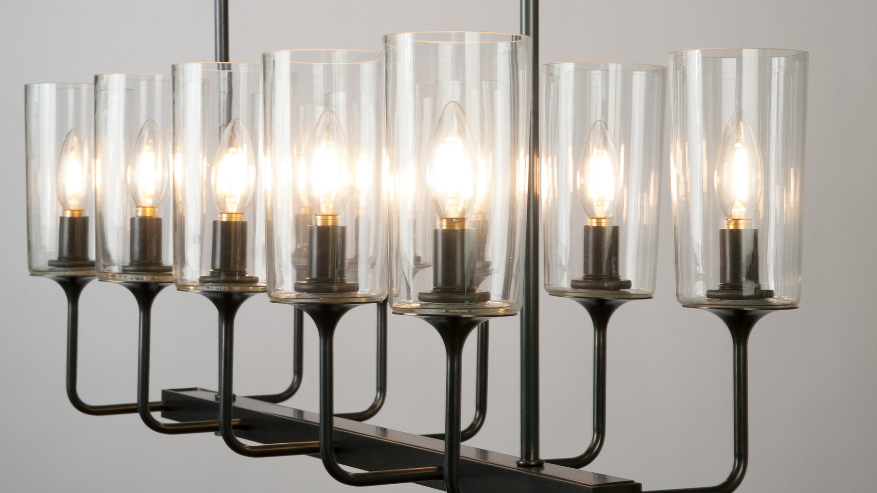 A close up of the Veronique 10 Linear Chandelier, and its ten evenly spaced arms that flare at the ends to hold clear glass shades, in a dark waxed bronze finish.