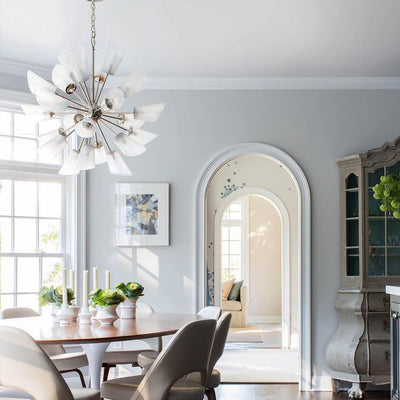 A polished nickel Vesta Chandelier, a round sputnik shape with blown opaline glass shades at the end of each arm, hangs in a sunny breakfast room with views out the windows and through multiple arched doorways. The space was designed by Carmina Roth.