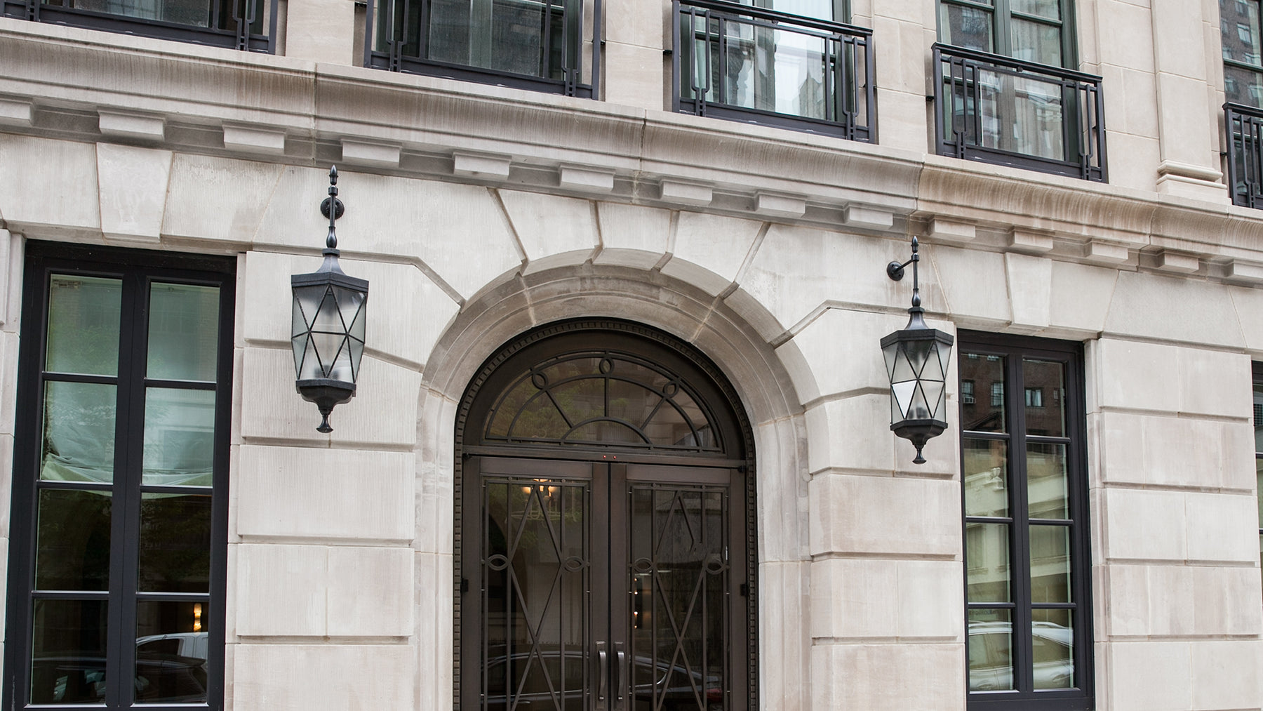A facing view of a pair of leaded glass and bronze exterior wall lanterns surrounding the limestone entry of an Upper East Side apartment building. The polyhedral lantern bodies consist of frosted glass lenses in a double hexagonal antiprism and are held between massive bronze castings.