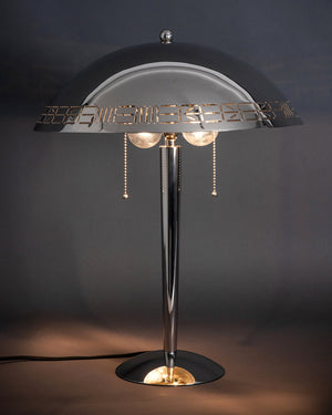 A view of the Commune Dome Table lamp with a pierced rim in a polished nickel finish, lit in a dark room against a gray backdrop.