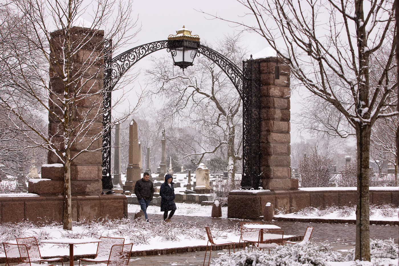 A large-scale custom outdoor lantern is the capstone for the archway at the entrance to a cemetery, pictured on a snowy day.