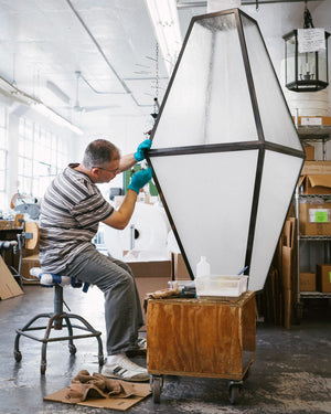 A Remains Lighting Co. staff member sits next to an eight-sided geometric lantern with brass framing and frosted seeded glass panels to put the finishing touches on its patina.