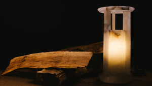 A cylindrical veined white alabaster table lamp with a pierced capital, supporting a broad top lens, with a pile a fire wood.