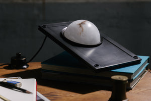 A table light made of a white alabaster globe set like a porthole in a rectangular dark waxed bronze plate sits on a table with books, pencils and papers.