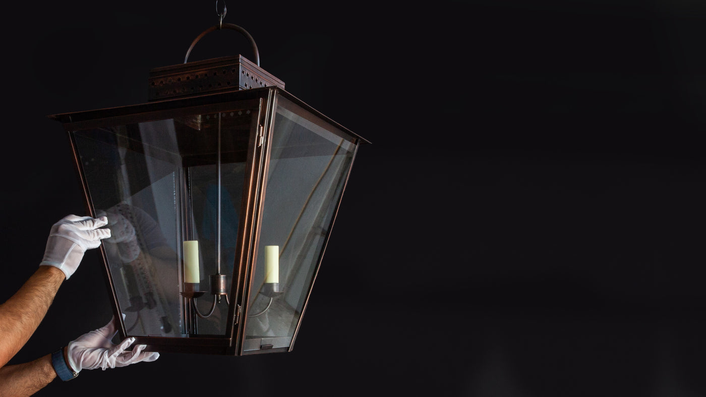 A pair of hands in white gloves puts the finishing touches on a four-sided antiqued copper ceiling lantern hanging in a darkened setting.