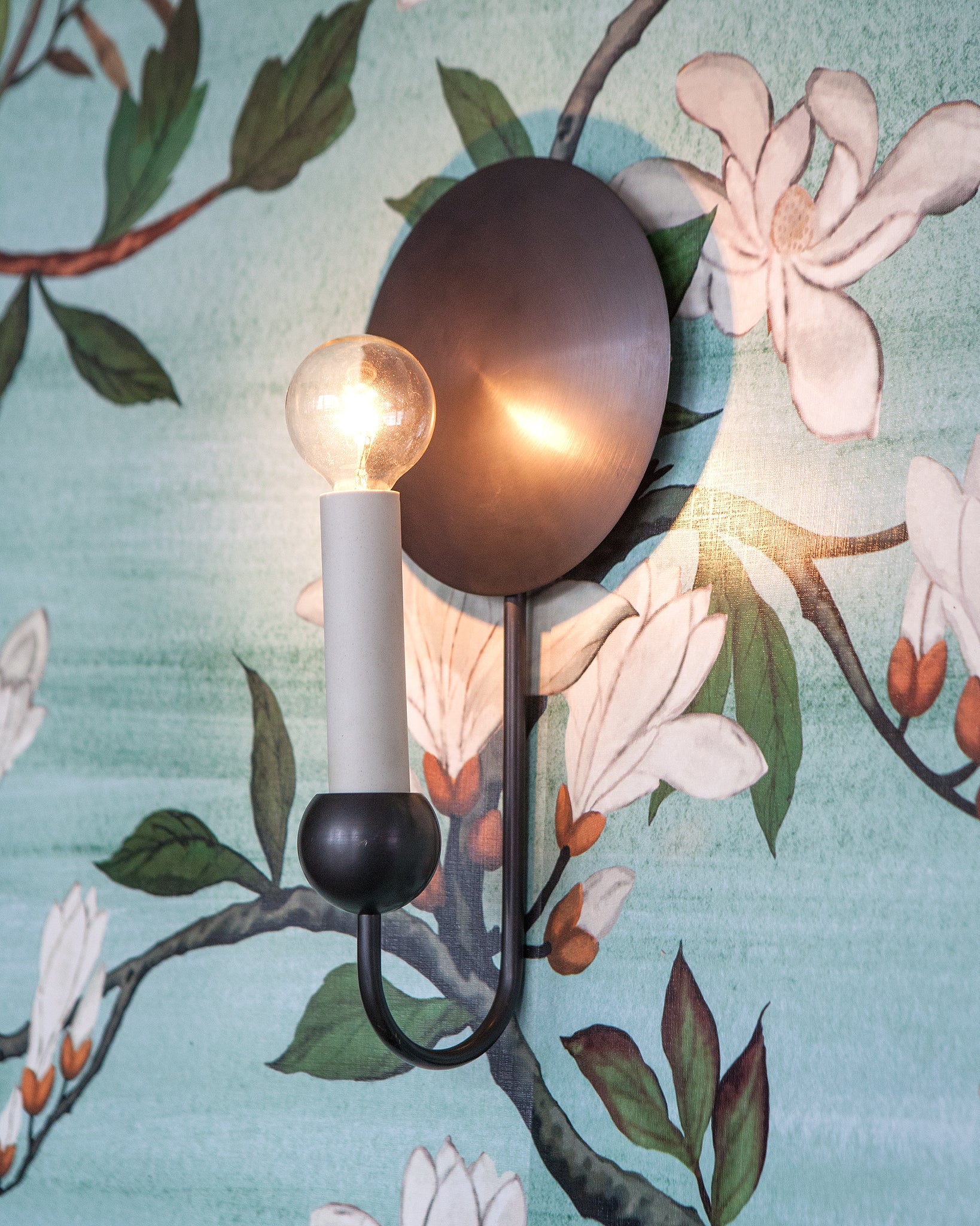 A wall sconce in antique brass with a gently domed backplate and spherical candle cup of solid brass is shown mounted and lit on a wall with pale blue wallpaper featuring magnolia blossoms.