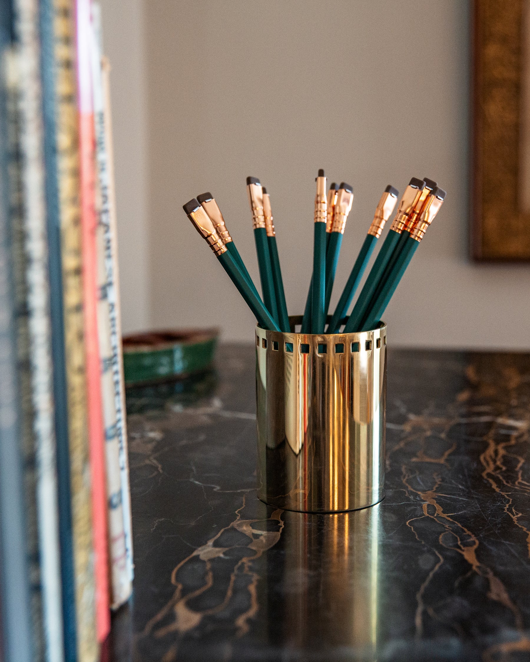 A polished brass Josef Cup by Remains Lighting Company filled with green pencils sits on a black marble surface with gold veining. A stack of books occupies the foreground on the left, slightly out of focus.
