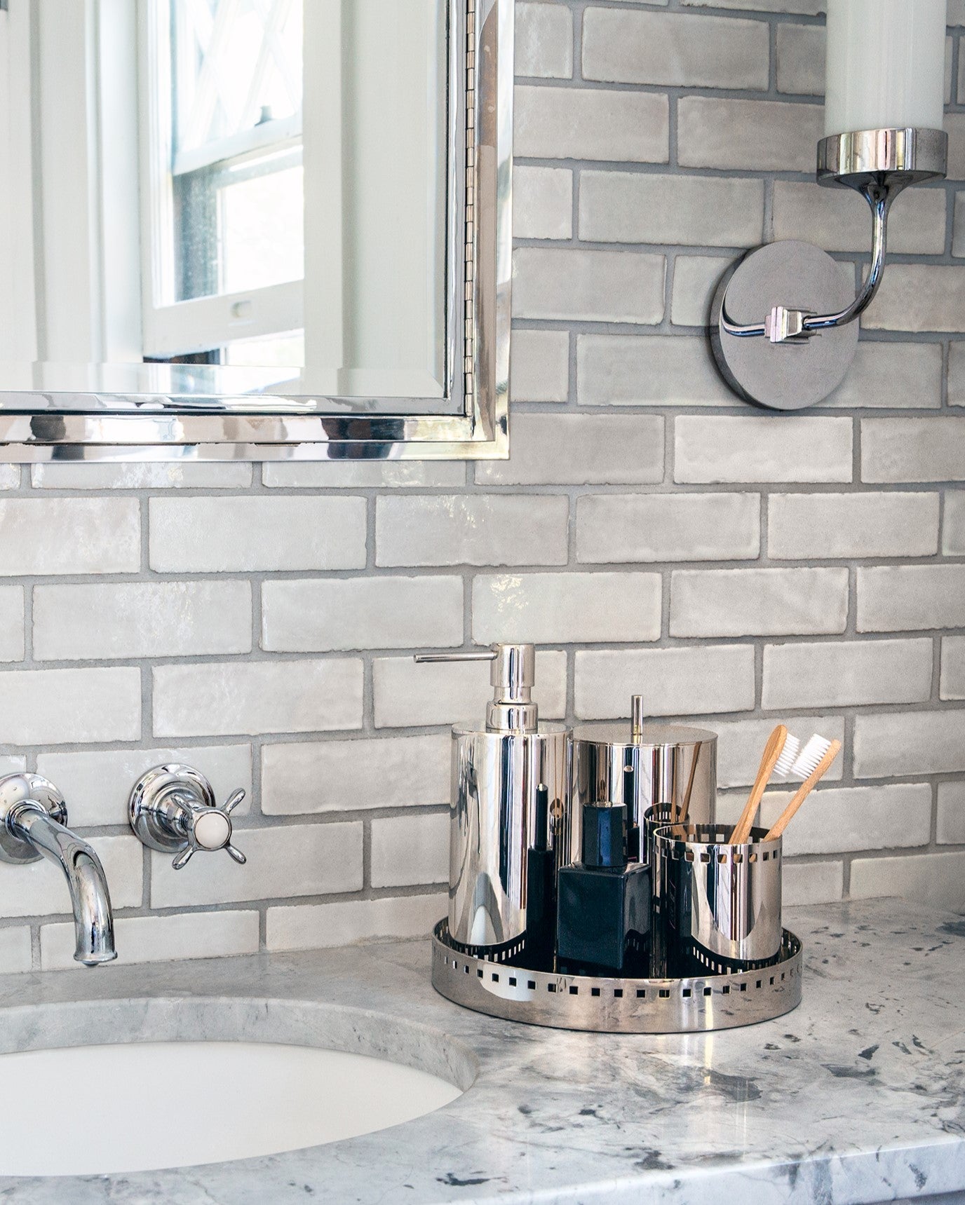 Josef Collection of Accessories in Polished Nickel on a marble bathroom sink. A sink, mirror, and sconce are installed on a grey tile wall. 