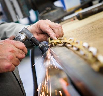 Sparks fly as chamfered bronze chain is hand hammered on a work bench.